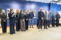 W jasno oświetlonym wnętrzu, z ciemnoniebieskimi ścianami, na tle plansz wywieszonych na ścianie stoi grupa kilkunastu osób, które kierują uwagę przed siebie.