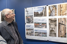 Mężczyzna w ciemnym garniturze, z uśmiechem, spogląda w kierunku zdjęć wywieszonych na ścianie.