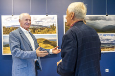 Dwóch mężczyzn z siwymi włosami rozmawia ze sobą. Uśmiechają się. W tle na granatowej ścianie zawieszone plansze z dużymi kolorowymi fotografiami.