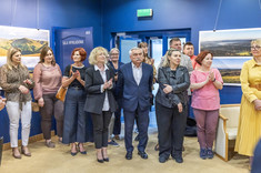 Ujęcie pomieszczenia, w którym stoi grupa kilkunastu osób. Wszyscy zwróceni w jednym kierunku, biją brawo i uśmiechają się. W tle na granatowej ścianie zawieszone plansze z dużymi kolorowymi fotografiami.