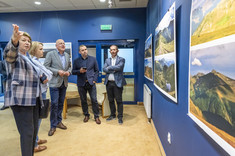 W pomieszczeniu kilka osób stoi przed prezentowanymi na ścianie fotografiami. Kobieta na pierwszym planie wskazuje coś ręka pozostałym rozmówcom.