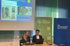 W lekko przyciemnionym pomieszczeniu po środku wysoko zawieszony jasny ekran, przed nim, przy stole siedzi dwóch prelegentów, mężczyzna i kobieta. W tle kilka wysokich na około 2 metry plansz w zielonym lub niebieskim kolorze z białymi napisami.