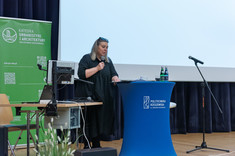 Kobieta ubrana na czarno stoi za pulpitem i przemawia do zebranych.  W tle duży biały ekran i zielony baner informacyjny z białym napisem - Katedra Urbanistyki i Architektury.