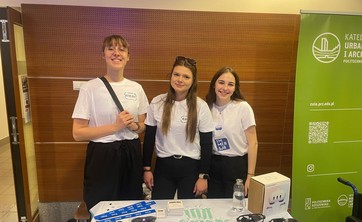 Na zdjęciu trzy uśmiechnięte studentki stoją za stołem przykrytym białym obrusem, na którym znajdują się ulotki, okulary i gadżety. Z prawej strony stoi zielony baner Katedry Urbanistyki i Architektury.