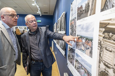 Mężczyzna w ciemnym garniturze wskazuje dłonią na fragment wywieszonego zdjęcia, a mężczyzna obok przygląda się fotografii.