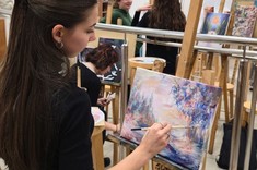 Ujęcie z boku. Kobieta z długimi ciemnymi włosami, ubrana na czarno maluje obraz ustawiony na sztaludze – drewnianym trójnogu.