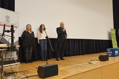 Na scenie znajduje się cztery osoby. Stoją na scenie w audytorium. Po lewej stronie stoi sprzęt komputerowy. W tle znajduje się duży ekran projekcyjny. Scena wykonana jest z jasnego drewna, a w tle widzimy ciemne kotary.