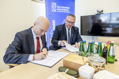 Na zdjęciu widać dwóch mężczyzn w garniturach, którzy podpisują dokumenty przy stole. Na stole znajdują się butelki wody mineralnej, filiżanka, pudełko oraz przekąski. W tle widoczny jest baner Politechniki Rzeszowskiej im. Ignacego Łukasiewicza oraz telewizor z kamerą. 
