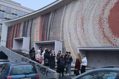 Na zdjęciu ściana budynku pokryta kolorową mozaiką. Przed wejściem do budynku na schodach zgromadzona grupa kilkunastu osób podczas luźnej rozmowy.