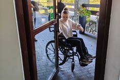Zdjęcie przed wejściem do budynku, podczas warsztatów projektowych. Osoba na wózku inwalidzkim próbuje sama otworzyć sobie drzwi uchylne i bez pomocy wjechać do budynku.