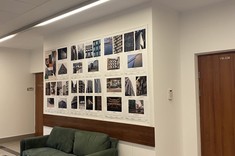 W pomieszczeniu na jasnej ścianie około dwudziestu zdjęć w stonowanych barwach rozplanowanych na prostokątnych planszach. Przed ścianą ustawione ciemno zielone kanapy.