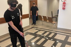 Na zdjęciu dwie osoby w opaskach na oczach poruszają się po korytarzu, używając białych lasek. Na podłodze widać wyznaczoną trasę z taśmy. W tle kilka krzeseł i drzwi.