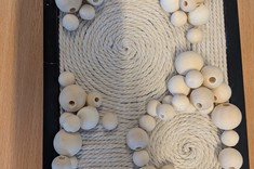 Na jasnym stole leży praca plastyczna. Na czarnej kwadratowej podstawie jasnym sznurkiem ukształtowane koła, linie, pomiędzy nimi linie z różnej wielkości koralików drewnianych.