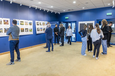 Wnętrze z ciemno niebieskimi ścianami. Na ścianach zawieszone białe plansze z kolorowymi zdjęciami. We wnętrzu, w kilku luźnych grupach, zgromadzone osoby odwiedzjące wystawę.