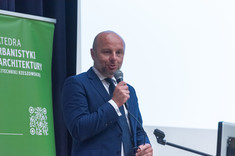 Mężczyzna w garniturze przemawia do zebranych. W tle biały ekran i zielony baner.