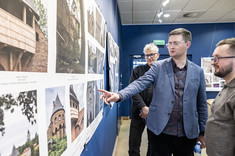 Mężczyzna w garniturze wskazuje dłonią na fragment wywieszonego zdjęcia, a dwóch mężczyzn obok przygląda się fotografii.
