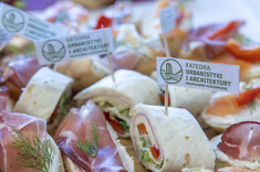 Zbliżenie na poczęstunek. Pośród jedzenia umieszone białe znaczniki z zielonym napisem: Katedra Urbanistyki i Architektury.