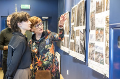 Dwie kobiety stoją i przyglądają się planszy ze zdjęciami, a jedna z nich wskazuje fragment zdjęcia.