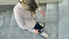 Kobieta w jasnym ubraniu, z ortezą na prawym kolanie, wchodzi po schodach, wspierając się na kuli.
