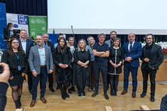 Grupa zgromadzonych kilkunastu osób pozujących do zdjęcia. Większość ubrana na czarno. W tle scena sali wykładowej.