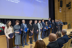 W pomieszczeniu przed sceną w rzędzie stoi grupa około dwudziestu nagrodzonych, oficjalnie ubranych osób.