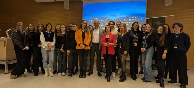 W pomieszczeniu stoi koło siebie około dwadzieścia osób i pozuje do zdjęcia grupowego. Większość ubrana na czarno. Po środku dwie osoby w kolorowych żakietach. W tle ekran z wyświetlonym zdjęciem.