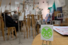 Zbliżenie na zieloną statuetkę. Na zielonej kwadratowej podstawie umocowany drewniany trójnóg, do którego przymocowano w pionie kwadratowy element: na białym tle zielona grafika, symbolizującą trzech ludzi w kaskach.