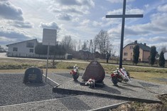 Zdjęcie przedstawia miejsce pamięci w plenerze, na którym znajduje się duży krzyż, tablica pamiątkowa oraz kamień oznaczony jako pierwsza stacja Drogi Krzyżowej.