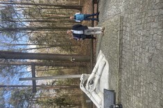 Zdjęcie przedstawia pomnik w lesie, który upamiętnia ofiary tragicznych wydarzeń. Na pierwszym planie widoczne są rzeźbione dłonie związane sznurem, symbolizujące cierpienie i zniewolenie. Obok znajduje się tablica z napisem „Pamięci ofiar”, a w tle widać drewniany krzyż oraz osoby stojące w pobliżu. Sceneria otoczona jest drzewami.