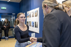 Mężczyzna ustawiony plecami do zdjęcia wręcza nagrodę młodej kobiecie.  W tle plansze ze zdjęciami.