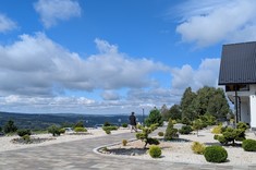 zdjęcie wykonane na zewnatrz budynku. Na pierwszym planie plac przy budynku z zadbanymi fragmentami zieleni. W tle naturalny krajobraz, zielone zalesione wzgórza i mocno niebieskie niebo.