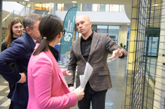 Na zdjęciu grupa 4 osób rozmawia ze sobą. Jeden z mężczyzn wskazuje dłonią na stojącą po prawej stronie pracę konkursową,