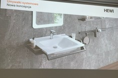 Na zdjęciu ekran z wyświetloną prezentacją. Na slajdzie widoczne wnętrze łazienki. Na tle szarej ściany biała umywalka z bocznymi uchwytami.