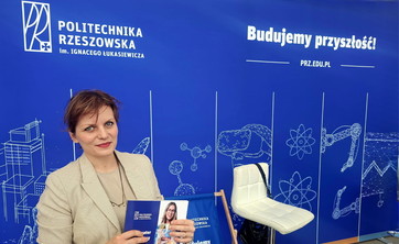 Na pierwszym planie kobieta w jasnej marynarce uśmiecha się do zdjęcia. W rękach trzyma folder reklamowy politechniki.  W tle ciemno ciemno niebieska ściana z białymi rysunkami i wyraźnymi napisami: Politechnika Rzeszowska i Budujemy przyszłość