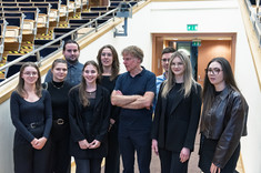 Zdjęcie grupowe grupy dziewięciu osób, kobiet i mężczyzn. Większość ubrana na czarno. W tle wyjście z sali wykładowej.