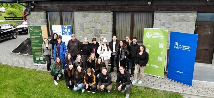 Zdjęcie wykonane z wysokości kilku metrów. Grupa około dwudziestu osób ubranych na sportowo pozuje do zdjęcia. Wszyscy spoglądają do góry. W tle kamienna ściana budynku.