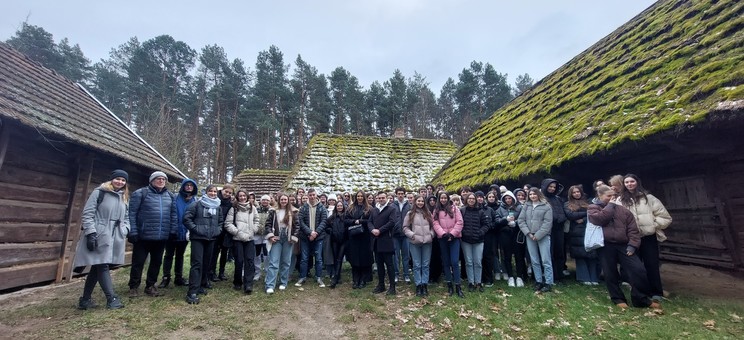 Pomiędzy dwoma drewnianymi chałupami z drewnianych bali z wyraźnie wystającym okapem i dachem pokrytym drewnianym gontem pokrytym częściowo mchem stoi grupa studentów wraz z prowadzącymi zajęcia. Grupa ponad trzydziestu osób. W tle widać ścianę wysokich drzew iglastych.