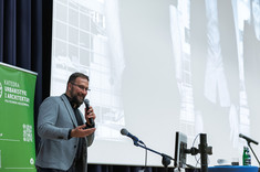 Mężczyzna w jasnej marynarce przemawia do zebranych.  W tle jasny ekran prezentacji i w narożniku zielony baner.