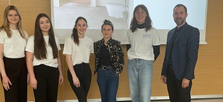 Na tle ekranu z wyświetloną prezentacją grupa sześciu osób. Po lewej mężczyzna w ciemnym ubraniu. Pośrodku grupy kobiet w białych podkoszulkach prelegentka w ciemnym ubraniu. Wszyscy się uśmiechają.