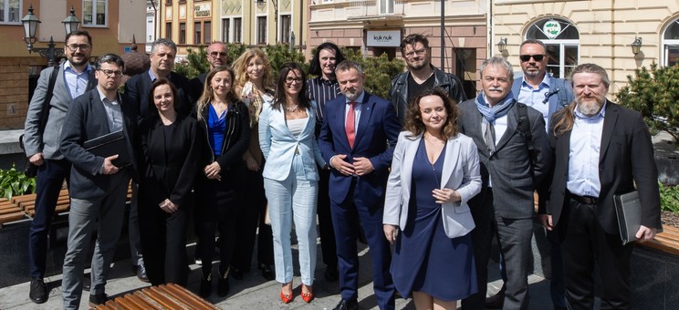Zdjęcie grupowe około dwudziestu osób, kobiet i mężczyzn, elegancko ubranych. W tle elewacje kamienic.
