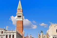 Zdjęcie zabytkowej architektury głównego placu z wyróżniającą się wysoką wieżą. Zdjęcie wykonane z przepływającego przed placem kanału z jaskrawo niebieską wodą.