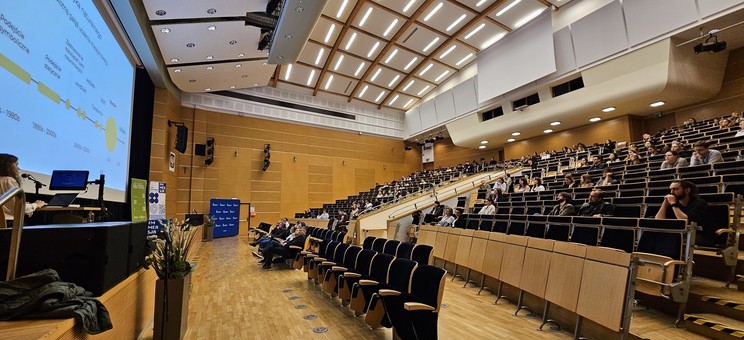 Zdjęcie przedstawia dużą aulę wykładową podczas trwania prezentacji. Po lewej stronie kadru widoczna jest prelegentka — kobieta z długimi, kręconymi włosami — stojąca za pulpitem z laptopem i mikrofonem. Na ekranie za nią widnieje slajd.  Widownia zajmuje większość kadru — sala jest wypełniona liczną grupą uczestników, którzy z zainteresowaniem słuchają wystąpienia.