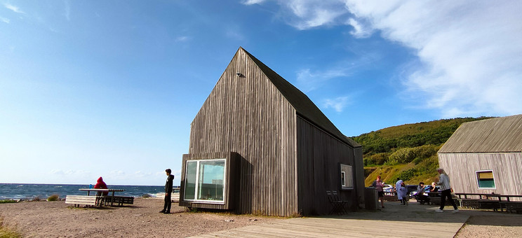 Zdjęcie: Pośrodku rozległej przestrzeni w naturalnym surowym krajobrazie zlokalizowany parterowy budynek. Bryła prosta z wysokim dachem, całość wykończona pionowym szarym deskowaniem. W tle zielone wzgórze i jaskrawo niebieskie niebo.