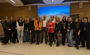 W pomieszczeniu stoi koło siebie około dwadzieścia osób i pozuje do zdjęcia grupowego. Większość ubrana na czarno. Po środku dwie osoby w kolorowych żakietach. W tle ekran z wyświetlonym zdjęciem.