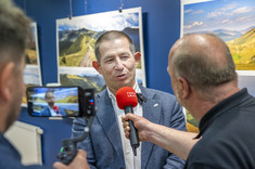 Mężczyzna, autor wystawy, w ciemnej marynarce udziela wywiadu dziennikarzowi z mikrofonem. Widoczne jest nagrywanie wywiadu na telefonie komórkowym. W tle na granatowej ścianie zawieszone plansze z dużymi kolorowymi fotografiami.