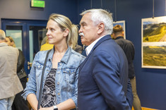 Na zdjęciu obok siebie stoi kobieta ubrana w niebieską kurtkę i mężczyzna w ciemnej marynarce. Rozmawiają ze sobą. W tle na granatowej ścianie zawieszone plansze z dużymi kolorowymi fotografiami.