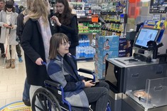 Zdjęcie przedstawia studentów w sklepie przy kasie samoobsługowej. Studentka na wózku inwalidzkim, w tle studenci w okularach oraz laskach. 