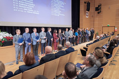 W pomieszczeniu przed sceną stoi w rzędzie grupa około dwudziestu nagrodzonych, oficjalnie ubranych osób.