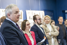 W jasno oświetlonym wnętrzu, z ciemnoniebieskimi ścianami, na tle plansz wywieszonych na ścianie stoi w rzędzie grupa elegancko ubranych osób. 