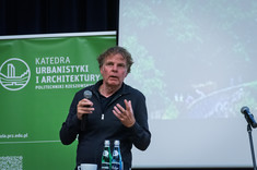 Mężczyzna ubrany na czarno prowadzi wykład. W tle jasny ekran prezentacji i w rogu zielony baner.